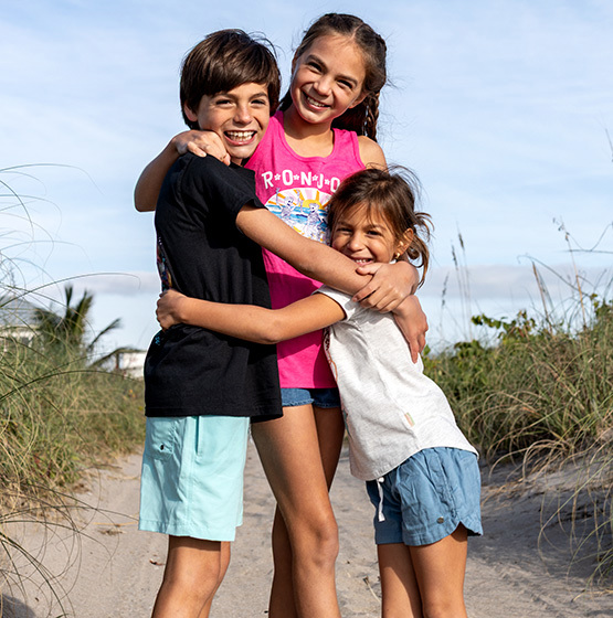 Three children stand on a sandy path near tall grass, smiling and hugging each other, with a blue sky in the background.