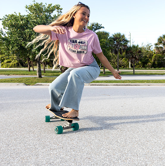 Woman with long hair rides a skateboard barefoot on a sunny street, wearing a pink graphic t-shirt and wide-leg jeans. Trees and a road are visible in the background.