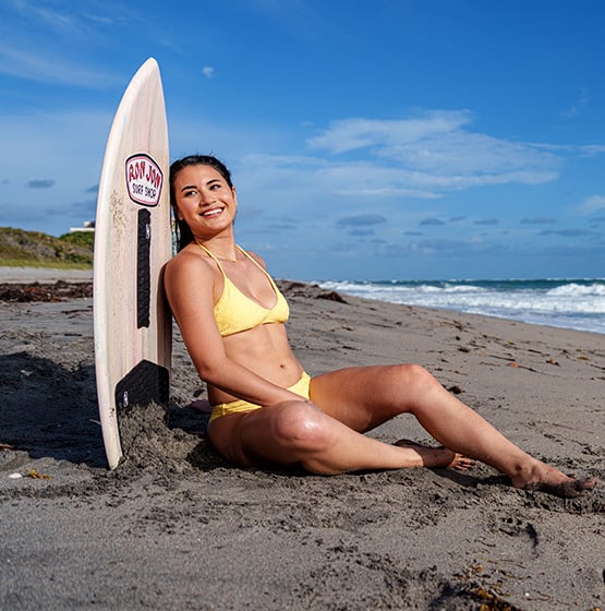 Woman in a yellow bikini sits on the sand next to a surfboard, smiling, with the ocean and blue sky in the background.