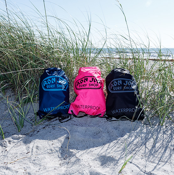 Three waterproof Ron Jon Surf Shop drawstring bags—blue, pink, and black—are placed on sand amid beach grass, with the ocean and sky in the background.