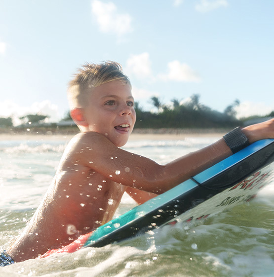 Young boy riding a bodyboard on ocean waves near a beach, with sunlight and palm trees in the background.