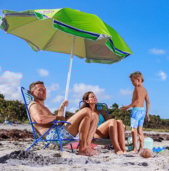 A family at the beach with a green umbrella. Two adults sit on chairs while a child stands nearby. They are surrounded by sand and beach gear under a clear blue sky.