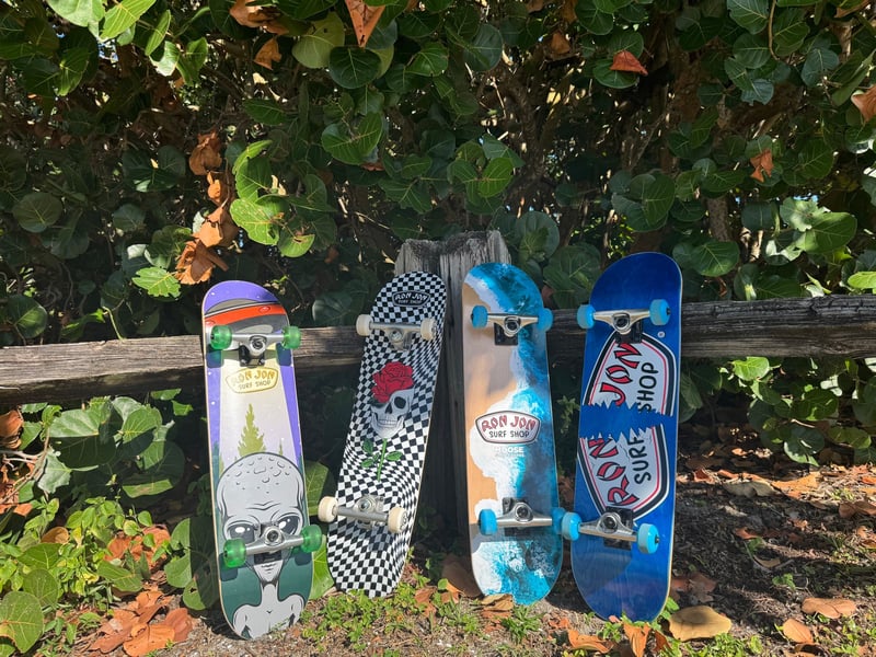 Four skateboards with colorful designs are leaning against a wooden fence in front of green foliage.