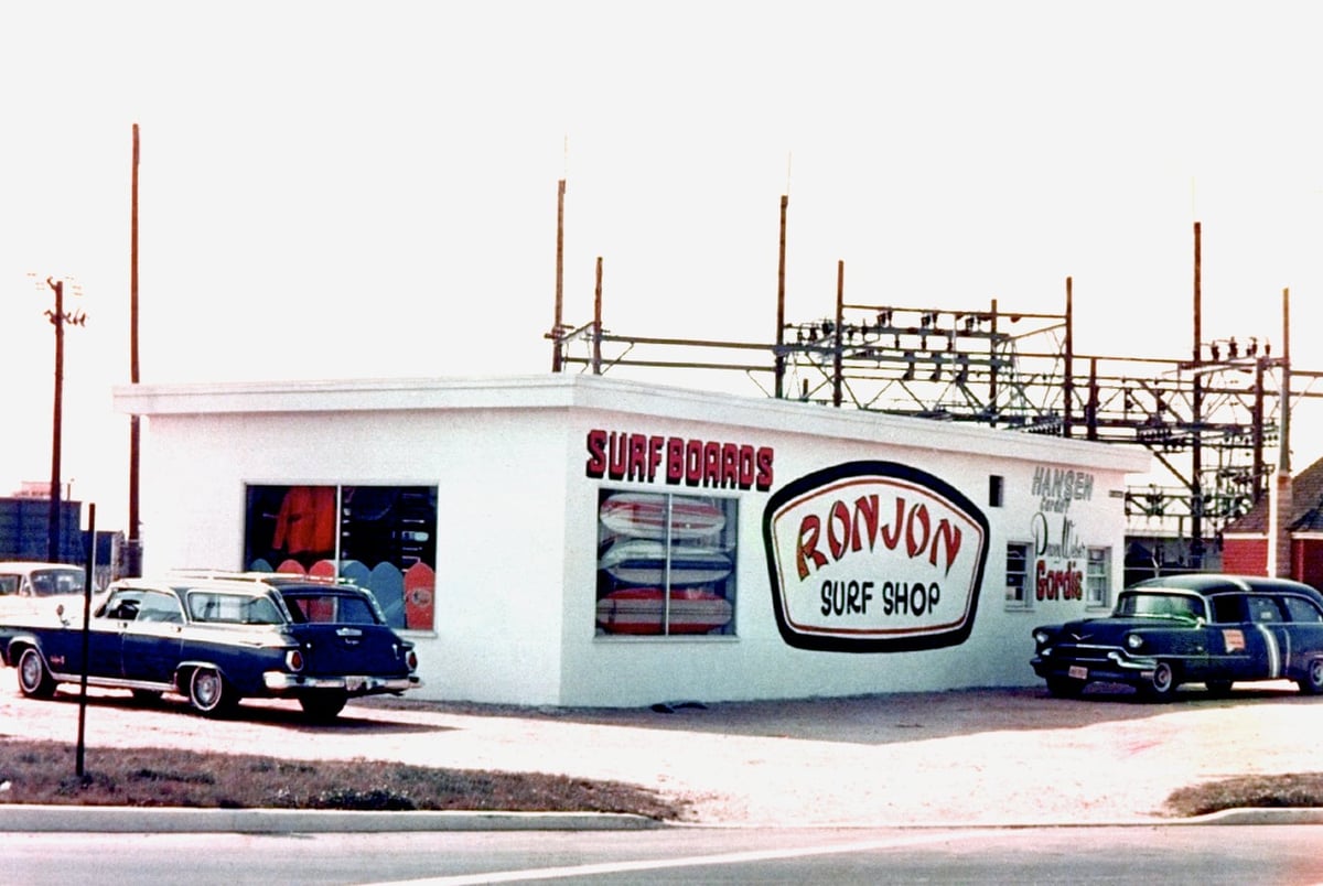 Vintage photo of Ron Jon Surf Shop showcasing surfboards, embraced by classic cars, with a power substation in the background—a true nod to the carefree beach lifestyle.