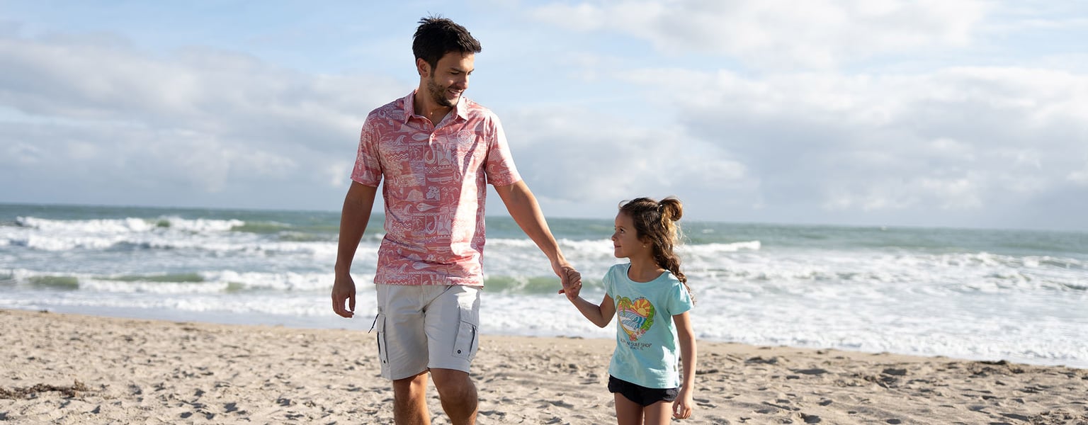 A man and a young girl walk hand in hand along a sandy beach with waves and a cloudy sky in the background.